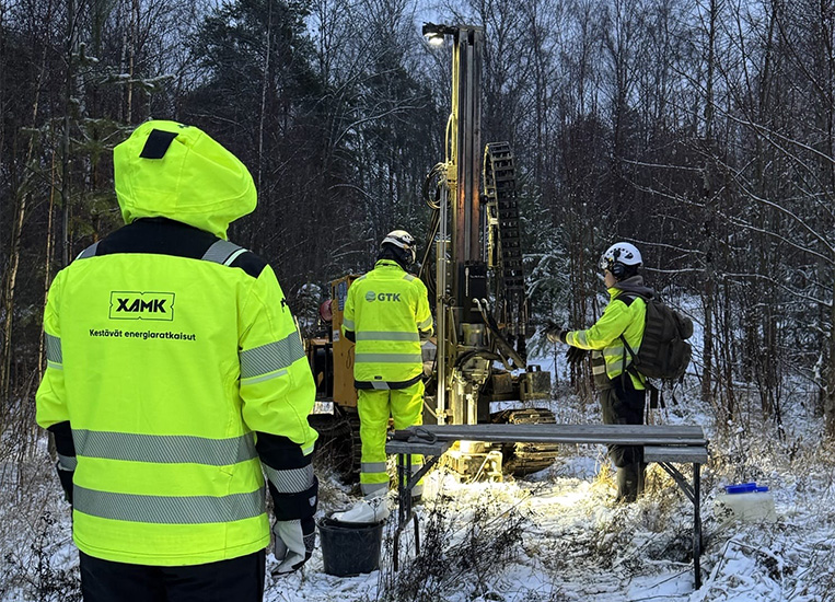 Kolme tutkijaa keltasissa huomiovarusteissa seisovat porauslaitteen ääressä lumisessa metsässä.