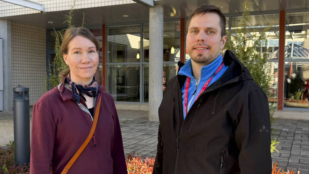 Mies ja nainen katsovat kameraan. Taustalla liikerakennuksen sisäänkäynti ja pensaita.