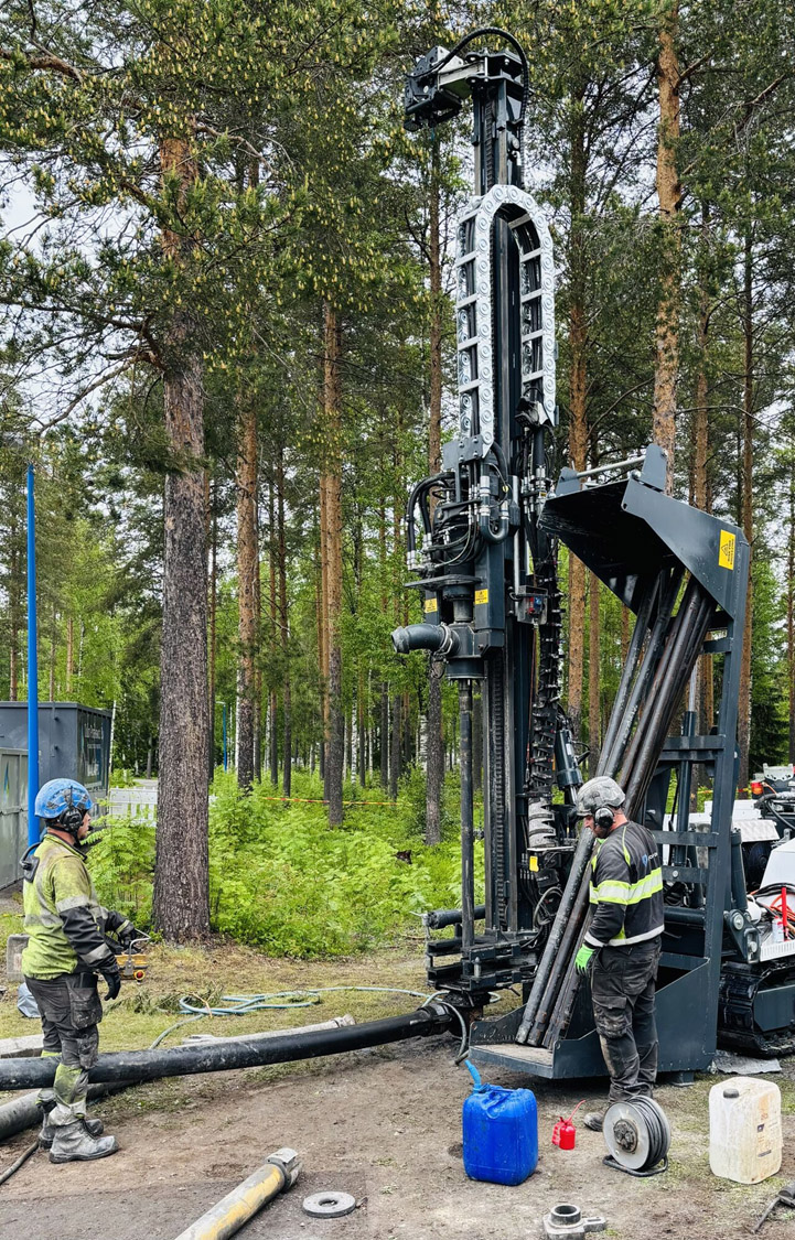 Isokokoinen porauslaite metsämaisemassa. Koneen äärellä kaksi turvavarusteisiin pukeutunutta henkilöä.